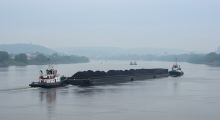 Black Coal Barge Being Pulled by a Tugboat Under a Misty Sky Across Calm River
