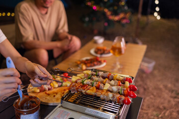 Friends enjoy a cozy outdoor evening barbecue with grilled skewers and drinks under string lights. One person grills food while another relaxes with a laptop, creating a mix of leisure and tech.