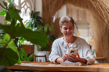 A cheerful senior woman enjoys a relaxing moment in a cozy, plant-filled cafe while using her smartphone. Surrounded by greenery, she smiles as she connects with others or reads online content.
