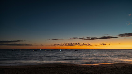 Evening after sunset by the ocean. The dark blue sky is highlighted with orange near the horizon. The clouds. Silhouettes of boats with lighted lanterns in the sea. Waves are foaming on a sandy beach