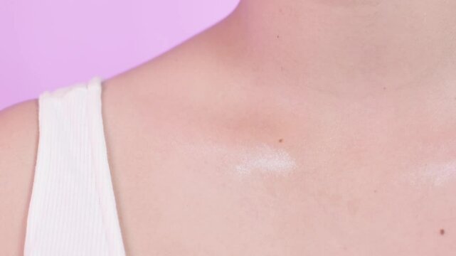 Close up of a woman applying highlighter on her collarbone with a fan brush, make up, cosmetics, on a pink background