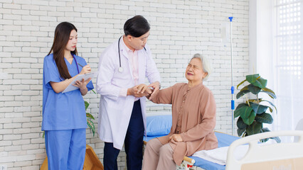 Fototapeta premium Asian senior woman sit on bed while consulting with doctor and caring nurse checking in hospital, Healthcare elderly patient and medical professional support muscle injury consultation wellness being.
