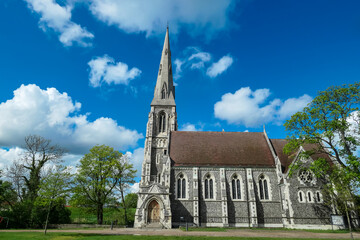 St. Alban's Church stands majestically, displaying its intricate Gothic Revival stone architecture and tall spire, surrounded by lush green trees and lawn in Churchillparken, Copenhagen under blue sky
