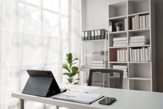 White desk in a contemporary office setting features a tablet. Business charts. A smartphone. And writing instruments. Bathed in natural light from a nearby window. Creating a productive workspace
