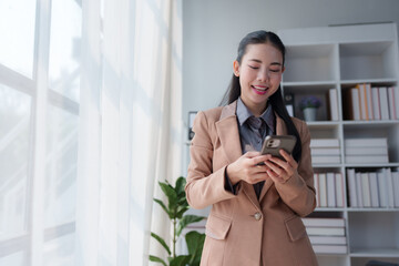 Fototapeta premium Young asian manager wearing a tailored suit, using a mobile phone in a modern office filled with bookshelves and natural light, smiling while checking messages on the screen