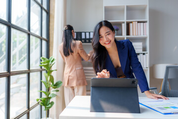 Young businesswoman smiling and using digital tablet while working on marketing plan in modern office with colleague arranging documents in background