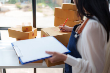 Online seller preparing packages for delivery. Carefully writing customer addresses on cardboard boxes while checking online orders listed on a clipboard. Managing logistics and inventory efficiently