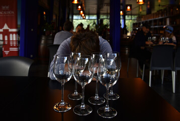 Person sitting in dark bar, head lowered onto table. Empty wine glasses in front of them