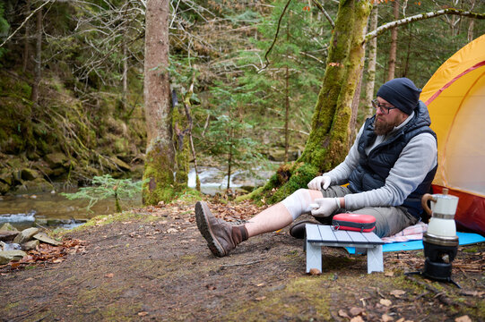 Hiker man with injured leg using first aid kit near the tent in the forest. A stressed trekker complaining alone in the mountain with broken knee. Traveler bandages leg after injury