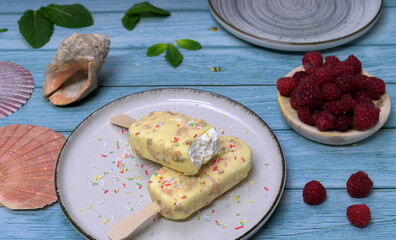 Homemade ice cream bars covered with white chocolate and red berries on a summery blue background, with raspberries and tropical decoration. Refreshing dessert concept
