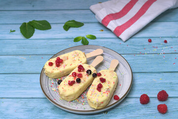 Homemade ice cream bars covered with white chocolate and red berries on a summery blue background, with raspberries and tropical decoration. Refreshing dessert concept