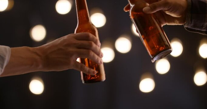 Closeup of male hands clinking beer bottles in festive atmosphere with warm bokeh lights celebration of friendship casual evening moment symbolic gesture of connection party cheers toast night vibe