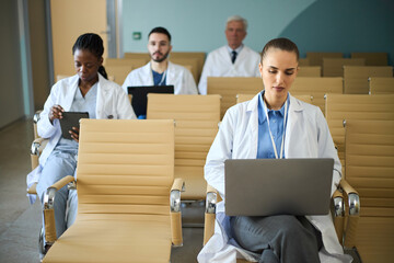 Group of medical professionals engaging with their laptops in hospital waiting room. Scene includes diverse team with Caucasian, African American doctor and senior practitioner