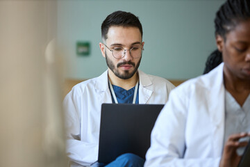 Medical professional wearing lab coat and glasses, concentrating on work while sitting in clinic with colleague nearby. Focused expression indicating dedication and attentiveness