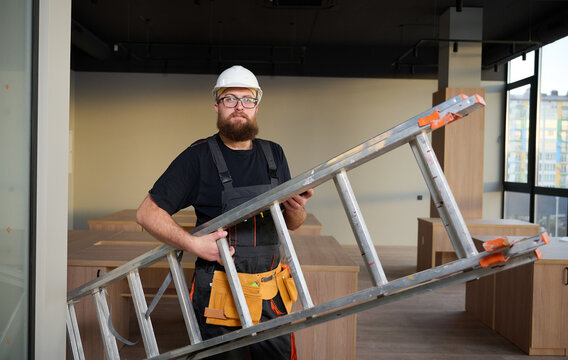 Construction worker holding ladder. Man carrying steel ladder. Safe at work. Bearded builder standing in warehouse wearing hardhat. Repairs concept. Construction builder worker carrying ladder