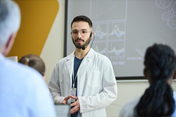 Medical professional in white lab coat engaging with audience during scientific presentation in modern conference room with projection screen