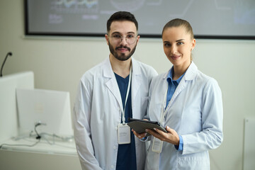 Portrait of two smiling healthcare professionals, one male and one female, wearing white lab coats, standing in clinic setting and holding tablet device while looking at camera