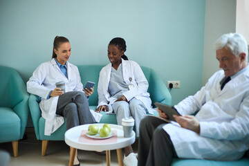 Obraz premium Medical professionals sitting in lounge, engaging in discussion over digital tablets and phones. Fruit and coffee cup placed on coffee table in front of them