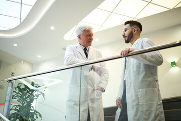 Senior doctor conversing with colleague in a contemporary hospital setting, both wearing lab coats and discussing medical advancements, with a plant and modern lighting in the background