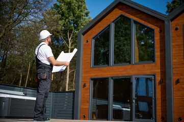 Architect hold plan. Bearded architect wearing hardhat holding project blueprints paper plans standing near comlex of new modern houses looking to something at construction site.
