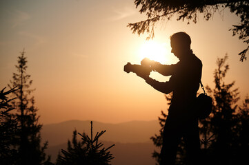 Silhouette of a photographer in the mountains taking pictures at dawn. Tourist holding camera on the mountain hill on sunrise background. Man takes photo of mountain in the rays of the setting sun.