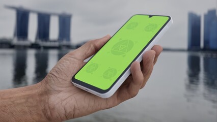 GREEN SCREEN CHROMA KEY Person holding a smartphone with a green screen near a waterfront, featuring a modern skyline and iconic buildings in cloudy daylight