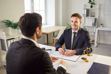 Smiling male lawyer in suit sitting at desk with legal documents, consulting client in modern office. Professional attorney listening attentively, providing advice and legal assistance during meeting.