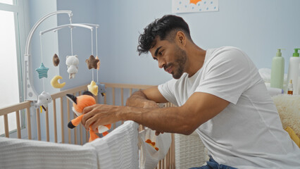 Young hispanic man with beard attentively playing with stuffed toys in a cozy bedroom, sitting by a baby's cradle, showcasing a warm and nurturing home environment.