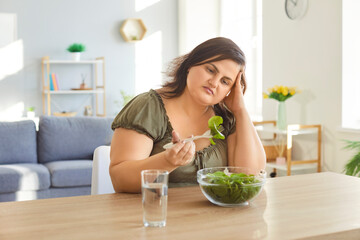 Overweight woman sitting at table in living room, looking at leaf of lettuce from bowl of fresh salad. Unhappy frustrated fat female eating vegetable salad, trying to adopt healthy eating habits.