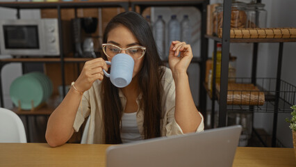 Woman sipping coffee while using a laptop in a cozy living room, surrounded by modern decor,...