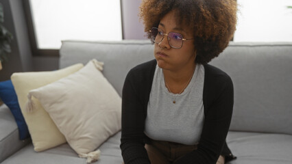 Woman sitting thoughtfully on a comfortable sofa in a cozy living room with pillows, wearing...