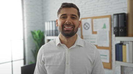 Young hispanic man smiling confidently in modern office interior with documents and plants in the background, showcasing a professional work environment.