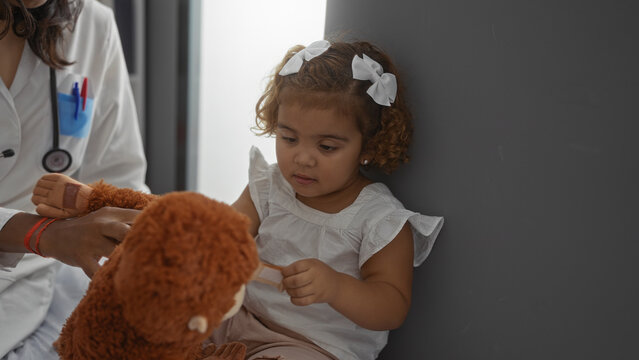 Pediatrician examining young girl with toy in hospital, woman in white coat using stethoscope, child holding teddy bear, clinic setting, indoor medical interaction