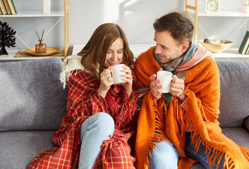 Smiling young couple sitting on sofa under woolen blankets in cozy living room and drinking hot tea. Happy freezing pair trying to warm up while enjoying each other company in cold weather at home.