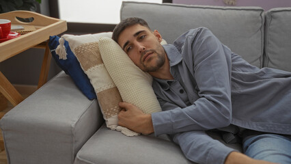 Fototapeta premium Handsome man relaxing indoors on a comfortable sofa at home, lying on pillows and gazing calmly ahead in a cozy living room setting.