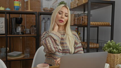 Woman feeling neck pain in cozy apartment kitchen, surrounded by homey decor, featuring a laptop and modern furnishings, radiating a casual and relaxed atmosphere.