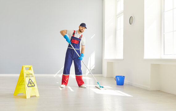 Male young bearded janitor in uniform washing floor with caution wet floor sign in empty room or at office. Man cleaner with mop and bucket mopping indoors. Cleaning service concept.