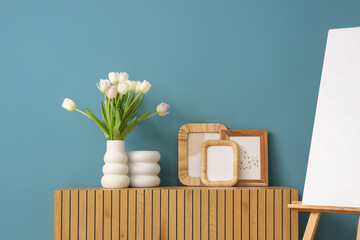 Photo frames and vase with tulips on commode near blue wall in room