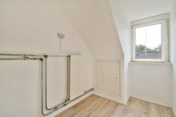 An interior view showcasing minimalistic design with white walls, wooden flooring, and a window providing natural light. Industrial piping adds a unique touch to the space.