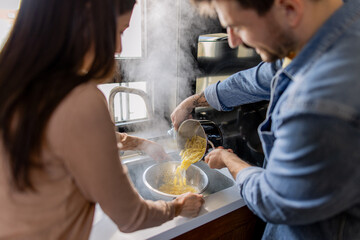 Man and woman draining steaming hot pasta in a colander, enjoying a moment of teamwork in their cozy kitchen sink