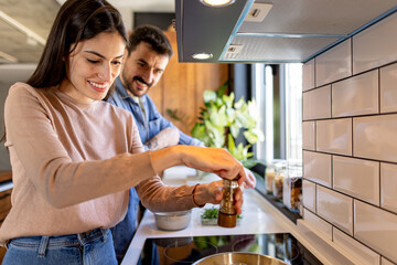 Happy couple preparing food together in their modern kitchen, adding spices and pepper to the meal