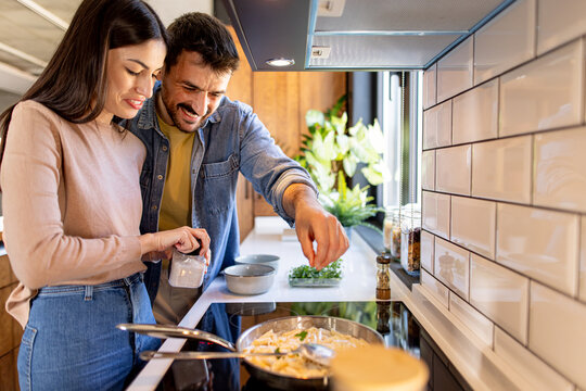 Young loving couple cooking together adding spices and salt in a pan with pasta in a modern kitchen - Powered by Adobe