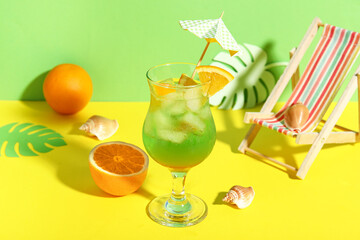 Glass of tasty cocktail with orange, umbrella, paper palm leaves, shells and deck chair on colorful background