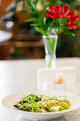  A fresh salad is beautifully arranged on a plate, featuring greens and cucumber. A vase with vibrant flowers and a cozy restaurant atmosphere enhance the inviting dining experience.
