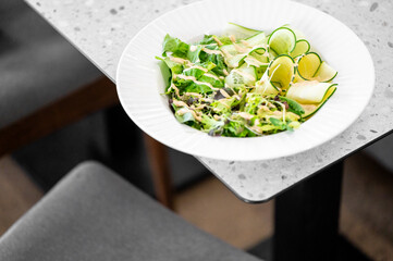 A close-up of a fresh salad featuring a mix of greens and cucumber slices, elegantly arranged in a white plate. The modern, minimalist table adds a stylish touch to the dining experience.