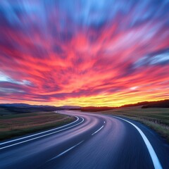Naklejka premium Curving road under a vibrant sunset sky with red, orange, and yellow hues. Landscape view with distant hills
