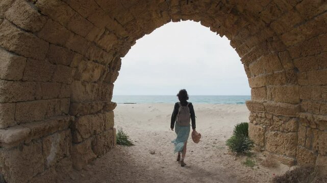 Travel and adventure. A female traveler passes through a stone arch of an ancient aqueduct and comes out to the sea.