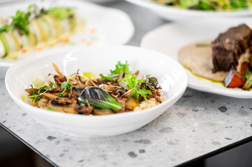 A beautifully plated dish of risotto with mushrooms and greens sits in the foreground, alongside other gourmet meals. The modern table setting enhances the elegant dining atmosphere.

