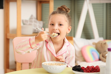 Cute little girl in pajamas eating cereals with yogurt at table in bedroom