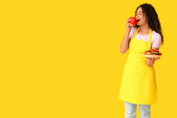 Beautiful young happy African-American woman with bell pepper and plate of grilled vegetables on yellow background